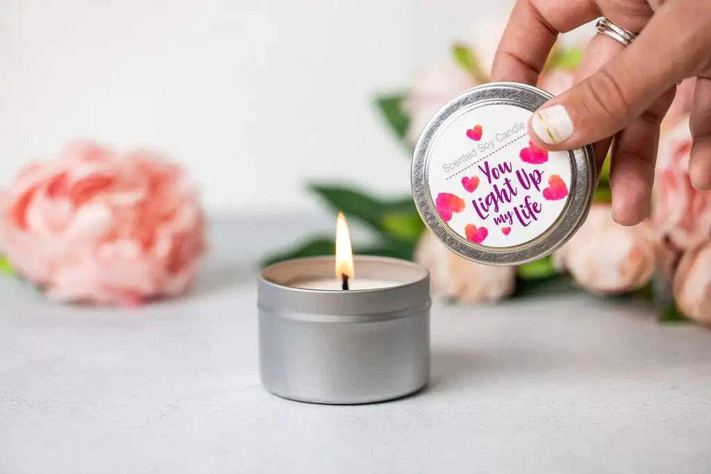 Hand holding lid of scented soy candle tin with "You light up my life" text next to lit candle and blurred pink flowers in background for Cherish Skincare Luxury Gift Box for Her