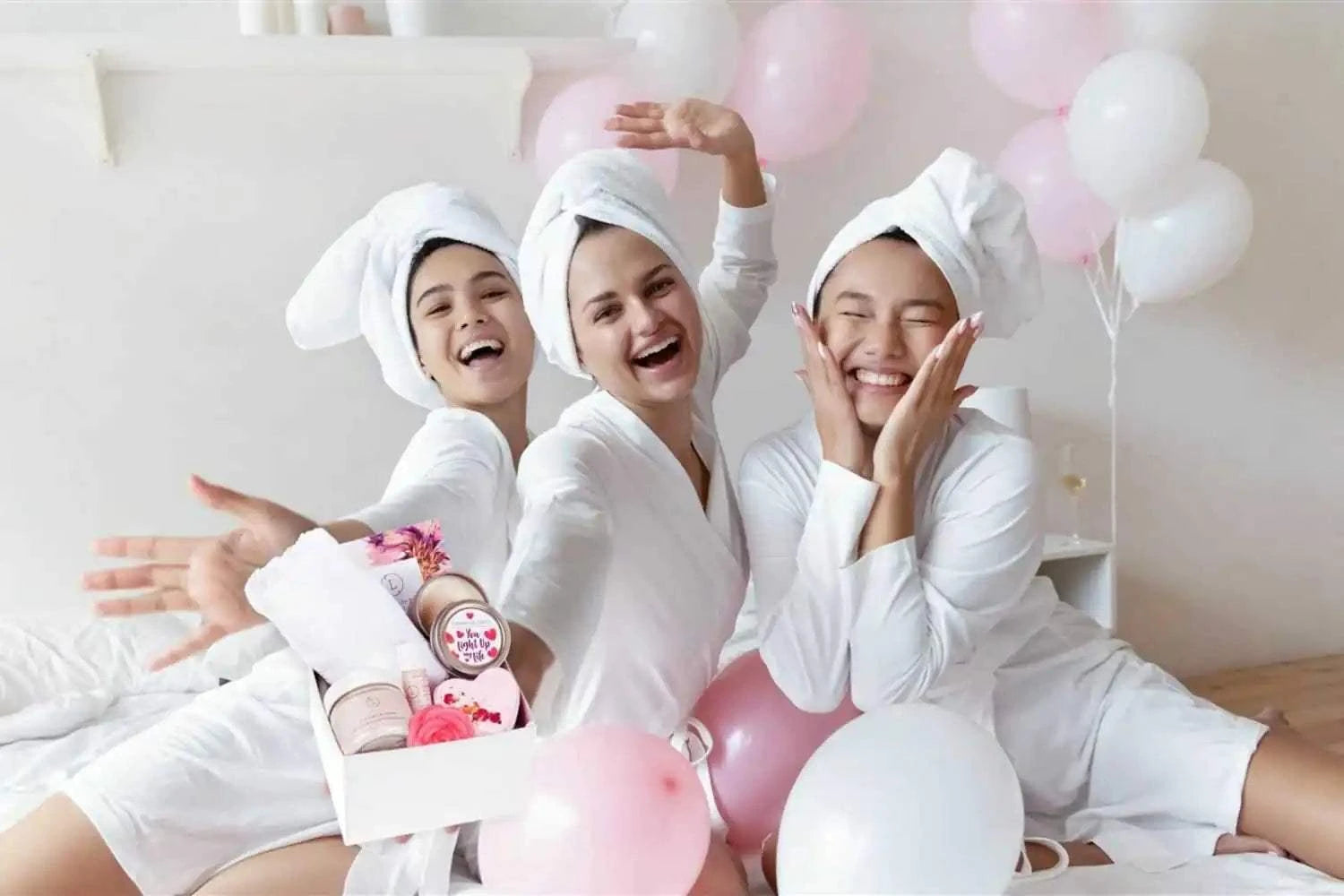 happy women in bathrobes with towels on heads holding Cherish Skincare Luxury Gift Box for Her with spa and skincare products and pink and white balloons